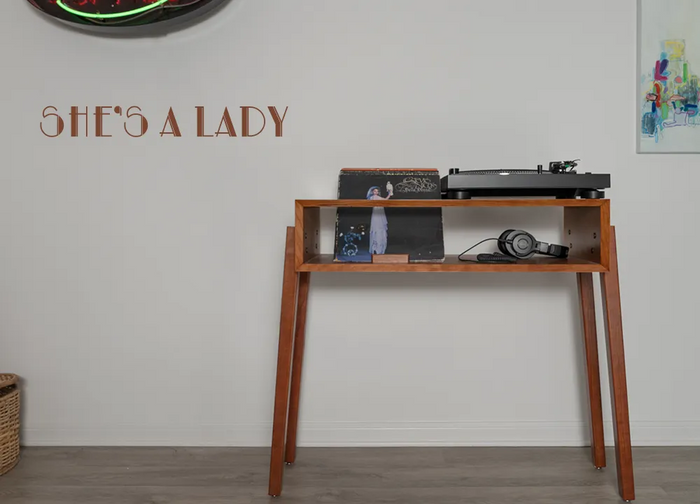 A handcrafted cherry record player stand with angled legs, featuring a turntable on top and vinyl records and headphones below. The words “She’s a Lady” appear on the wall beside it.