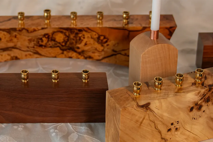 A display of handcrafted wooden menorahs in various shapes and wood types, featuring brass candle cups and natural burl grain