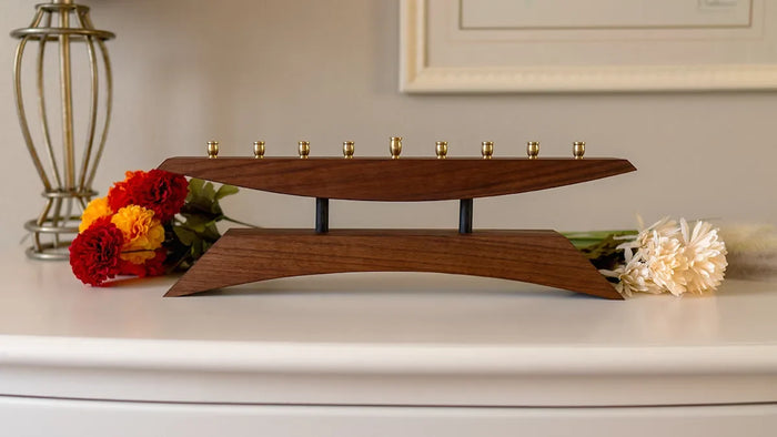 A modern walnut menorah with brass candle cups sits on a white surface, flanked by red, yellow, and cream flowers, blending sculptural form with handcrafted warmth.