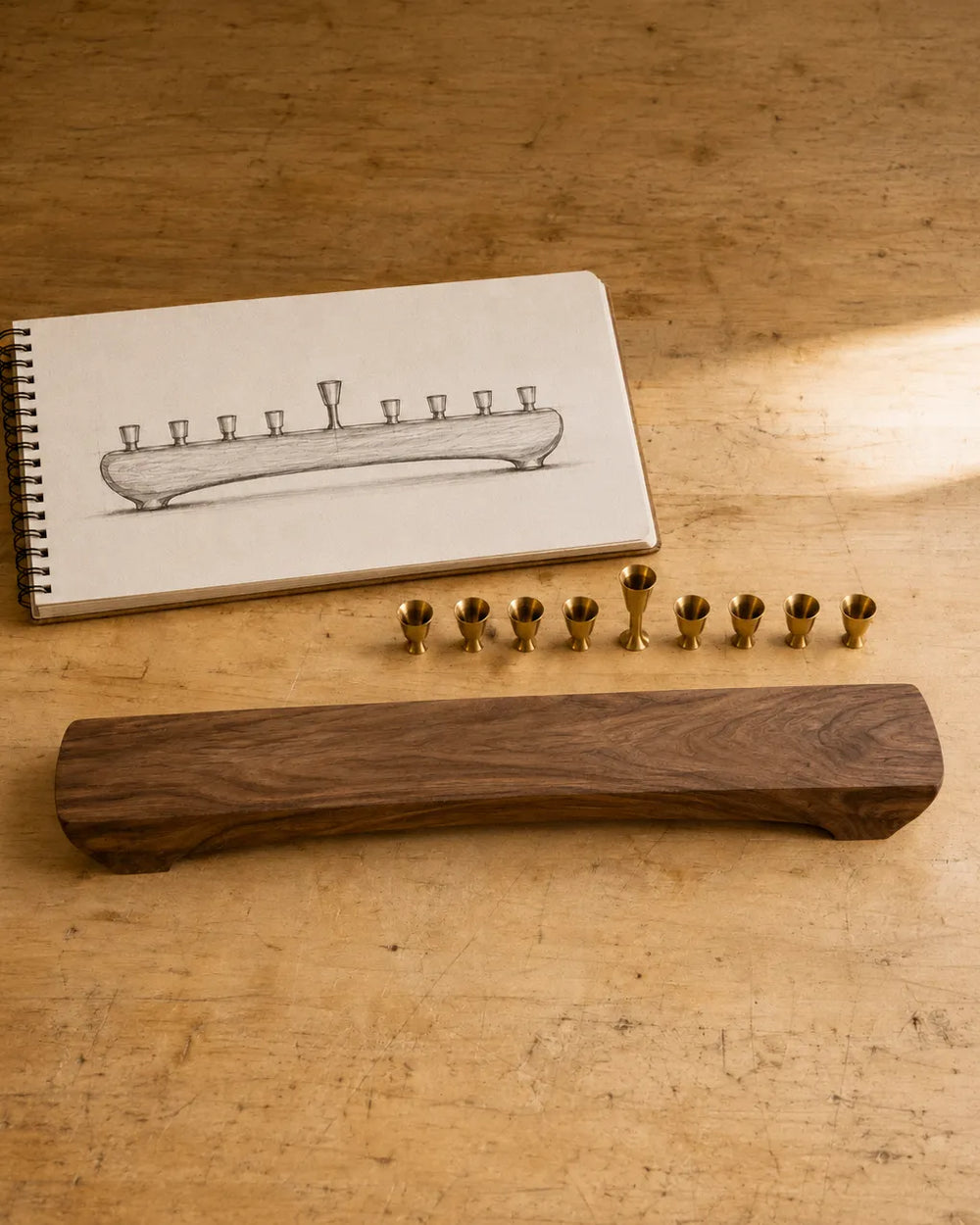 A pencil sketch of a nine-light menorah beside nine brass candle cups and a shaped walnut body on the bench
