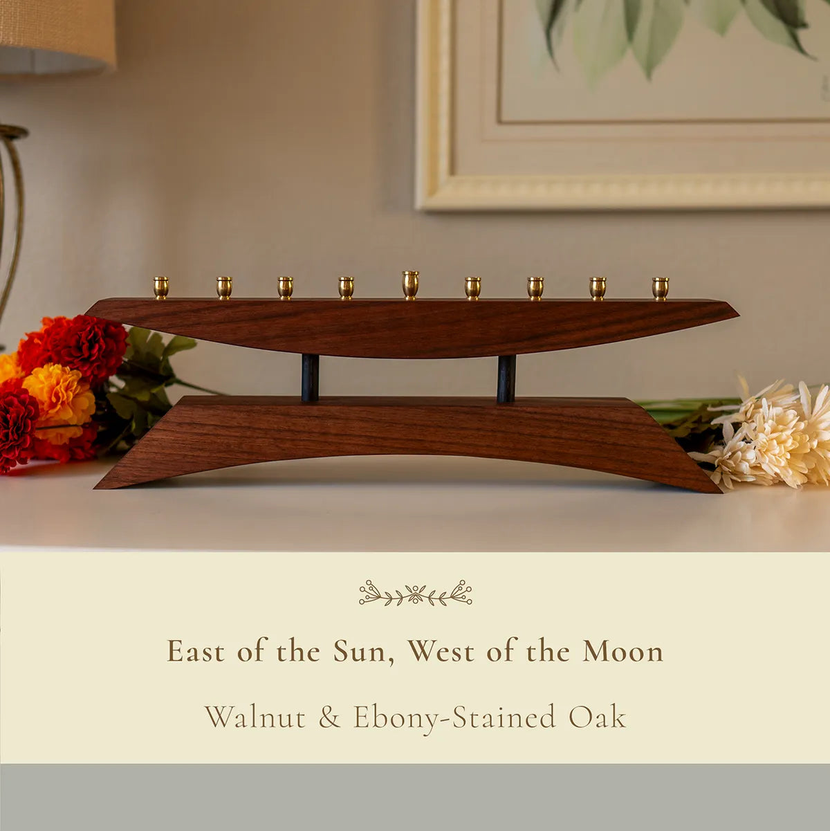 Modern wooden menorah titled “East of the Sun, West of the Moon,” handcrafted from walnut and ebony-stained oak, displayed with floral accents on a white mantle.