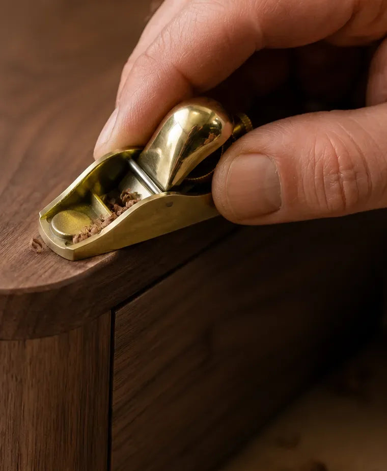 A hand easing the edge of a walnut top with a small brass block plane