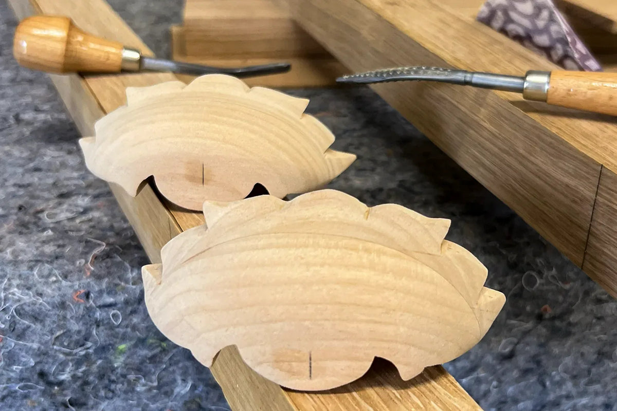 Two newly carved wooden bin pulls resting on the bench beside carving gouges