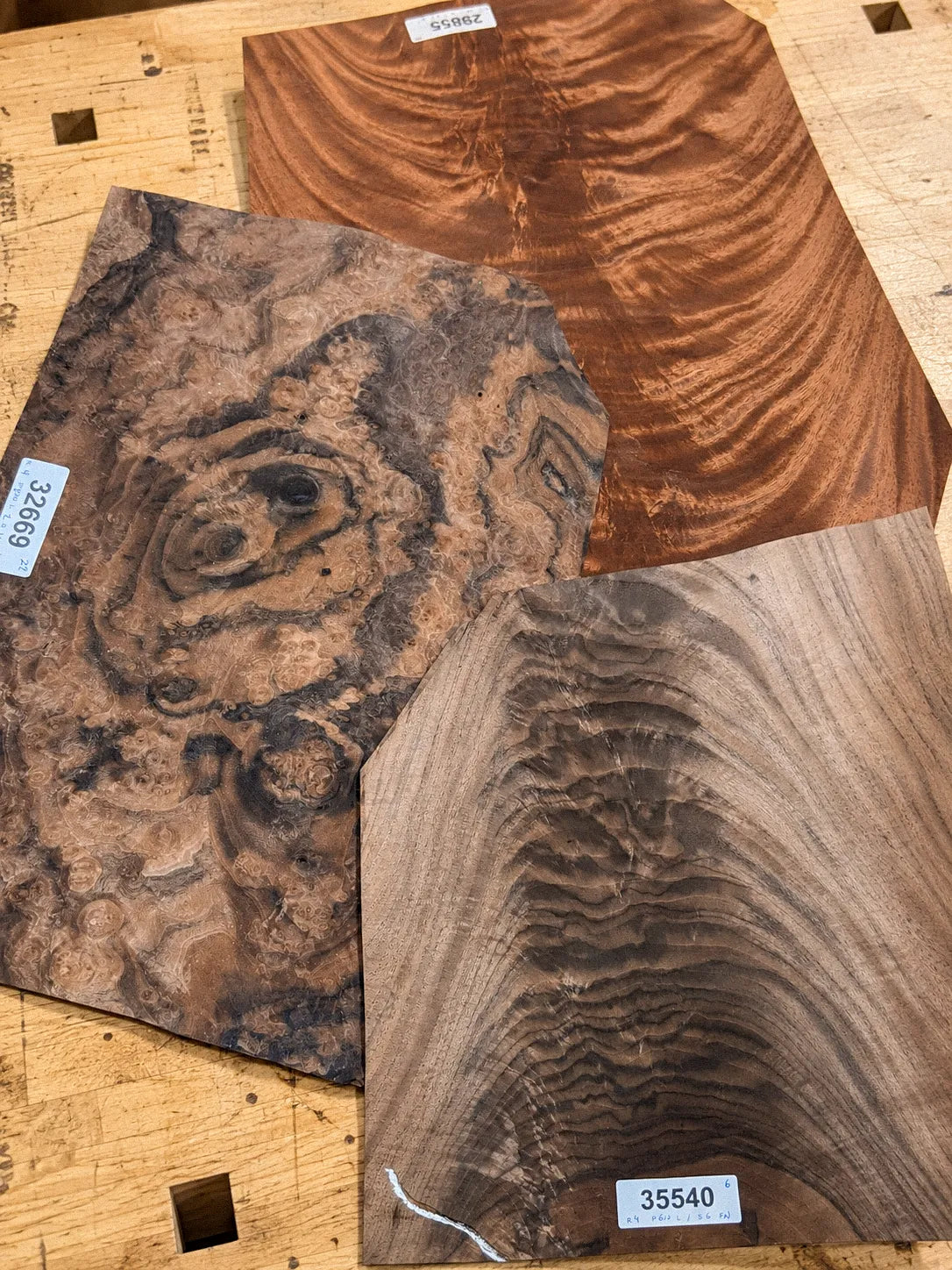 Three burl veneer sheets laid out on the studio workbench for selection, each with its supplier lot tag, showing the range of figure and grain across different burls