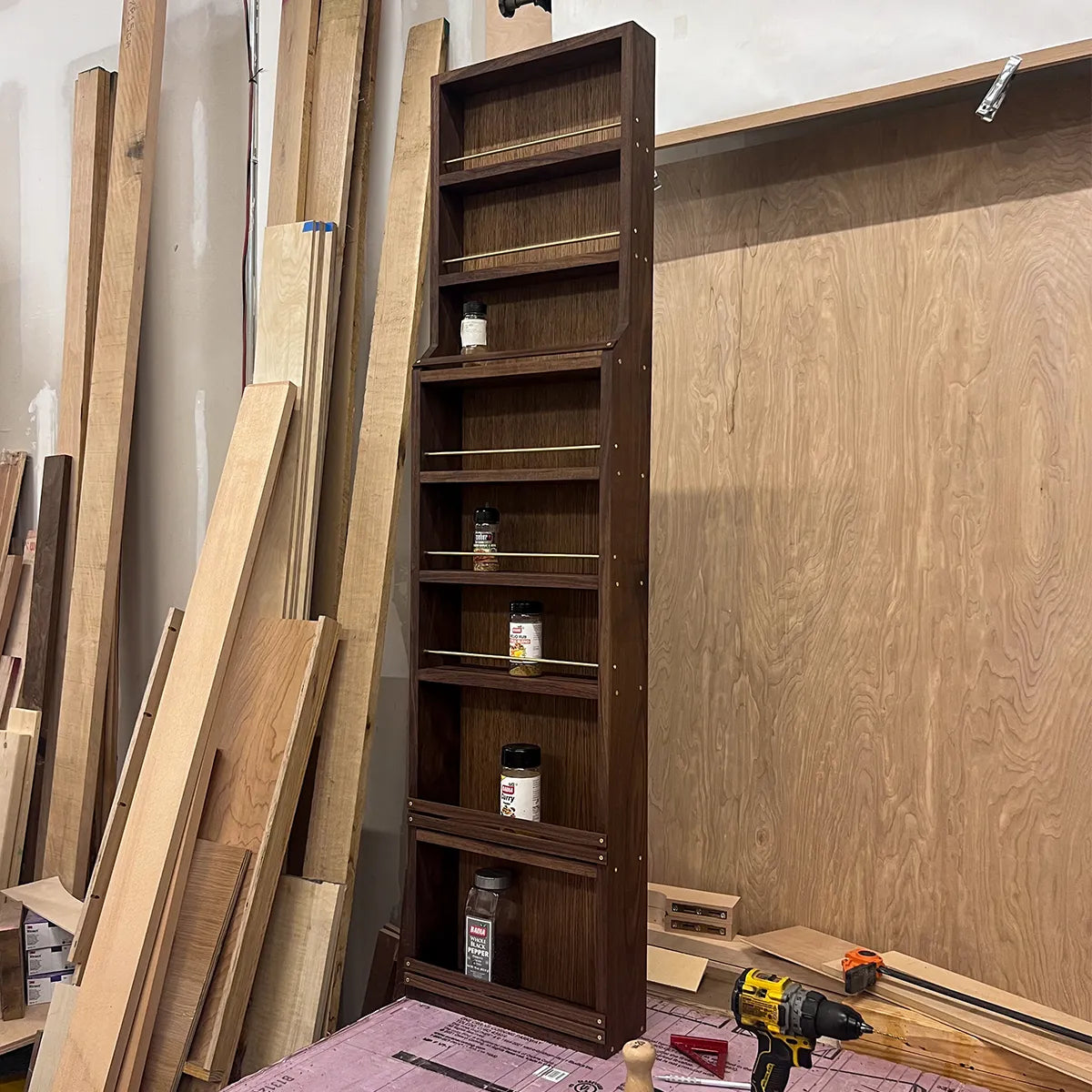 Tall walnut spice rack standing in the workshop, showing spice jars, brass rails, and its stepped-in mid-century design.