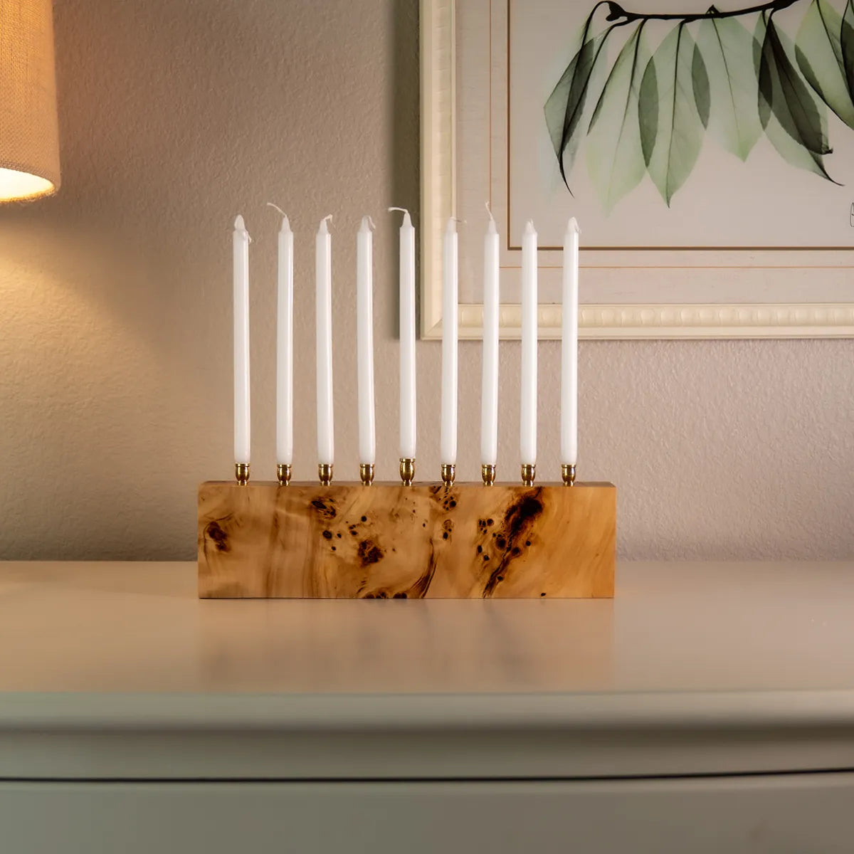 Solid mappa burl menorah with brass candle cups and white Hanukkah candles, displayed on a table beneath framed art.
