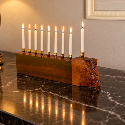 A unique live edge menorah with nine lit white candles, set on a dark marble countertop against a beige wall. The holder has a rich wood grain finish with a natural live edge on one end.